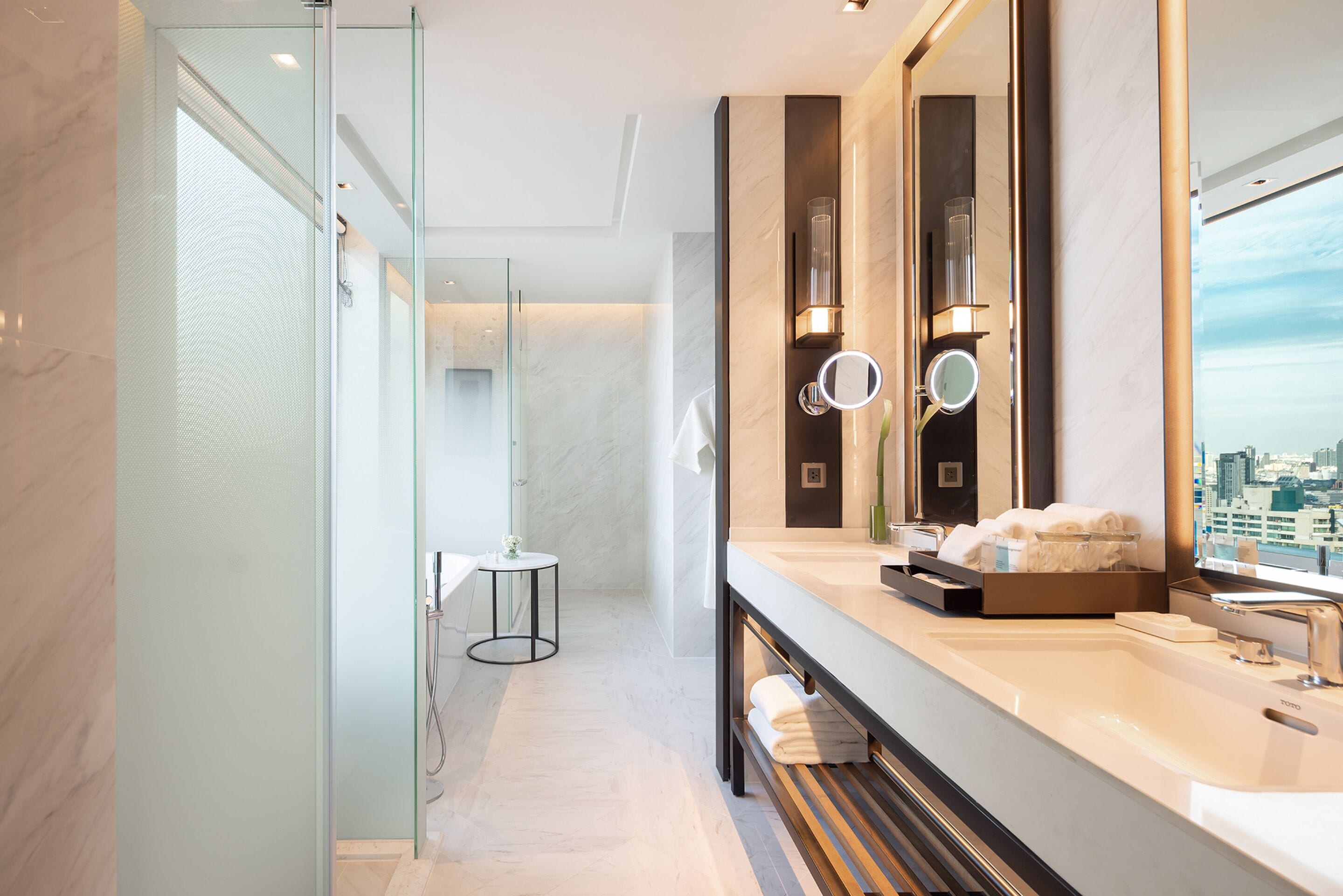 A modern bathroom with a glass-enclosed shower, a large mirror, and a city view.