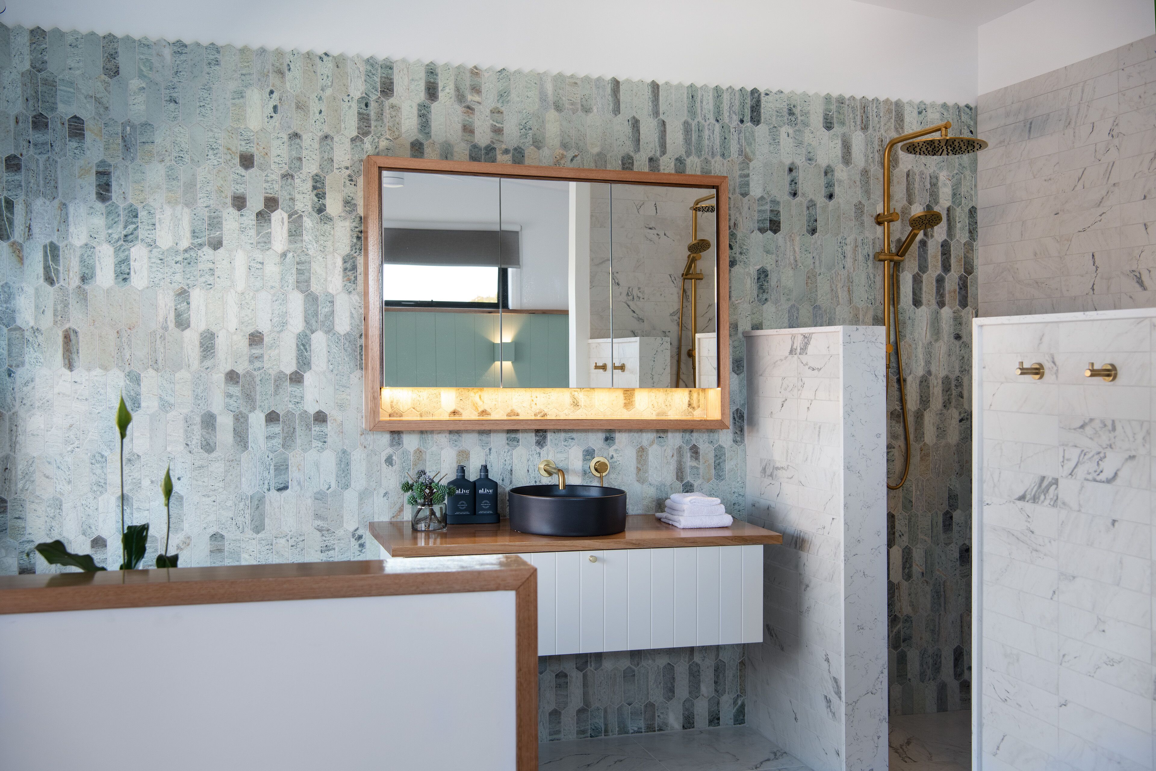 A bathroom with a stone-patterned wall, a wooden vanity, a large mirror, and a gold showerhead.