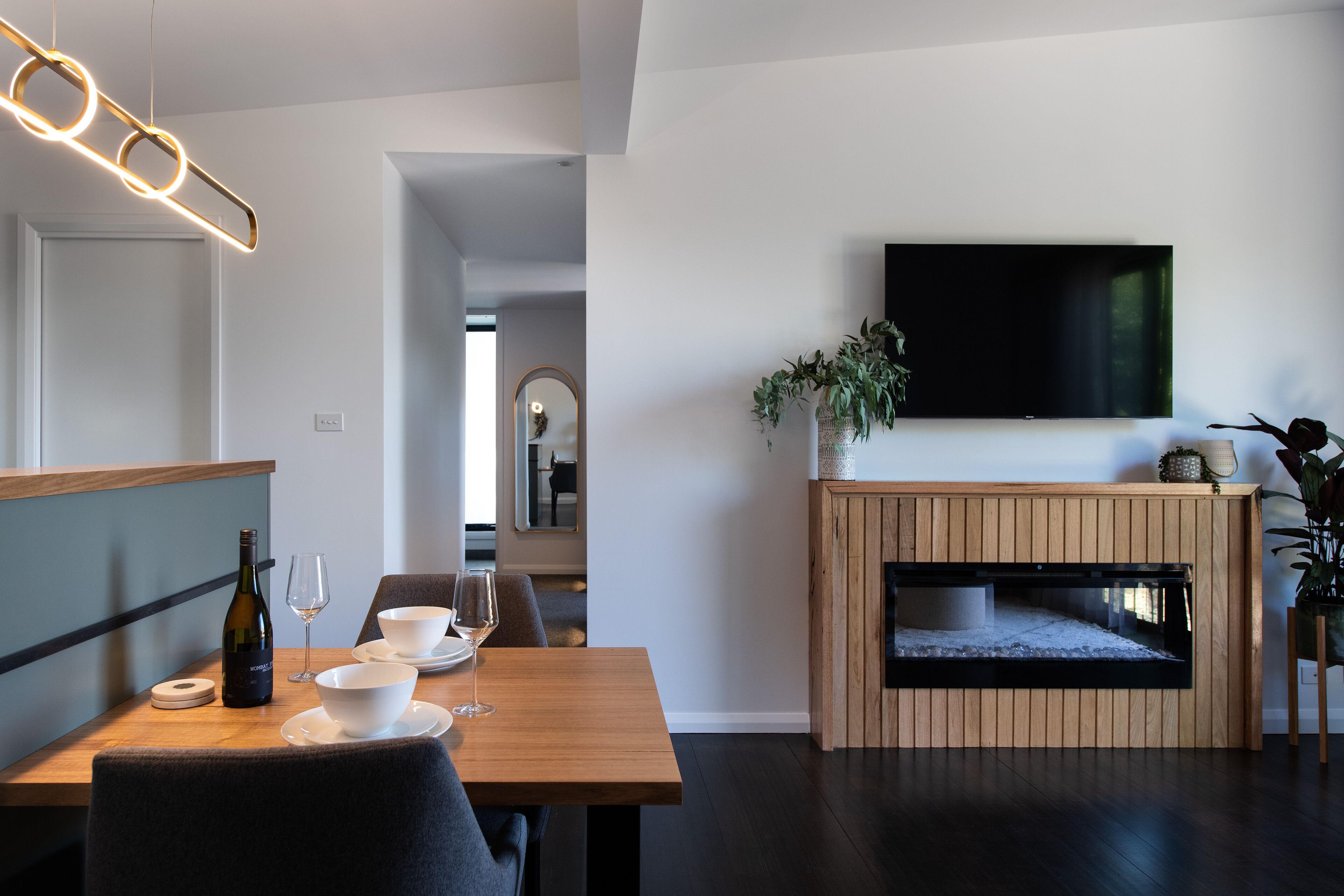A modern dining area with a wooden table, white bowls, wine glasses, and a bottle of wine. There is a flat-screen TV mounted on the wall above a wooden fireplace.