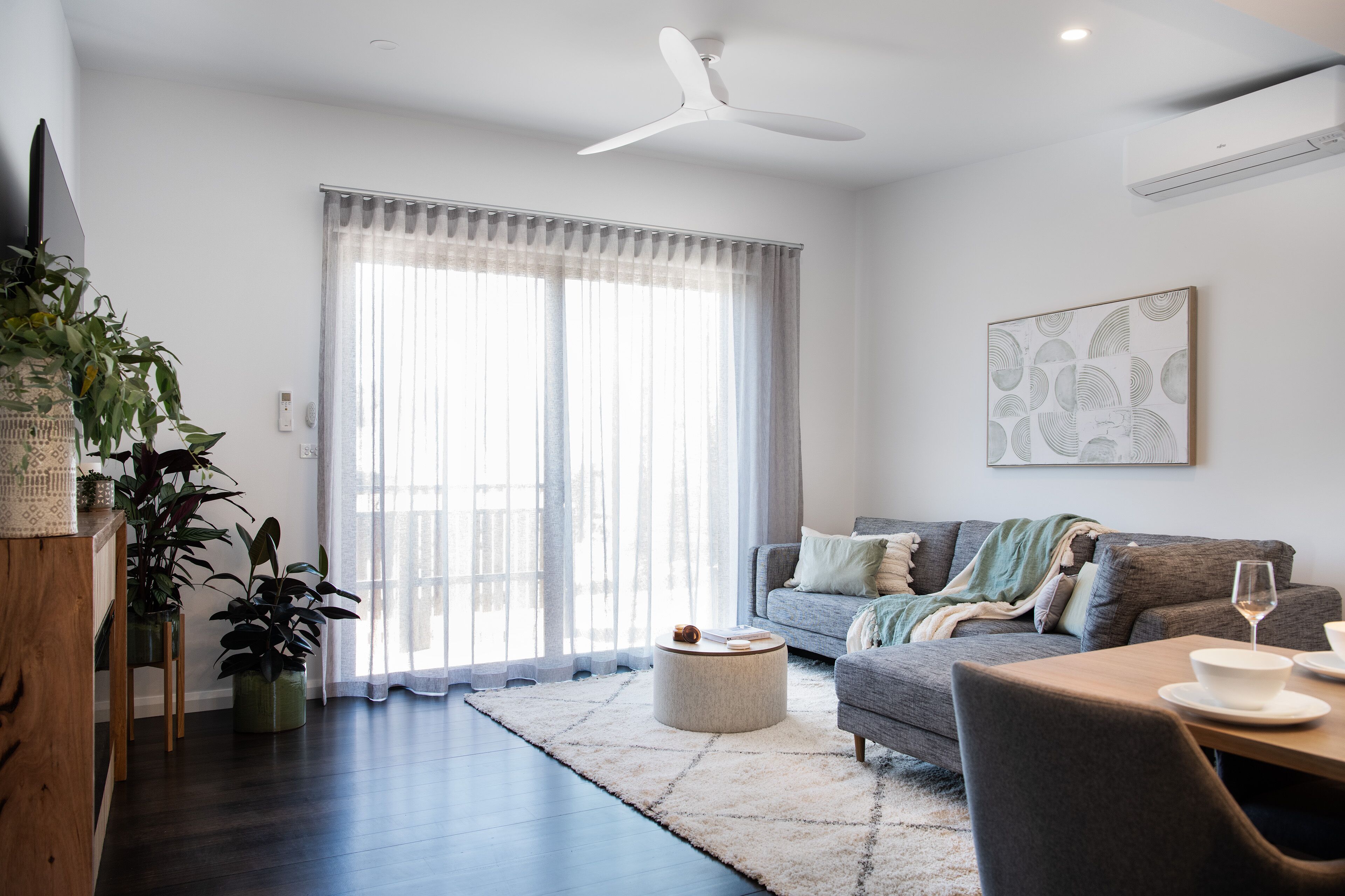 A modern living room with a sofa, coffee table, and dining area. There is a large window with sheer curtains, a ceiling fan, and an air conditioning unit.