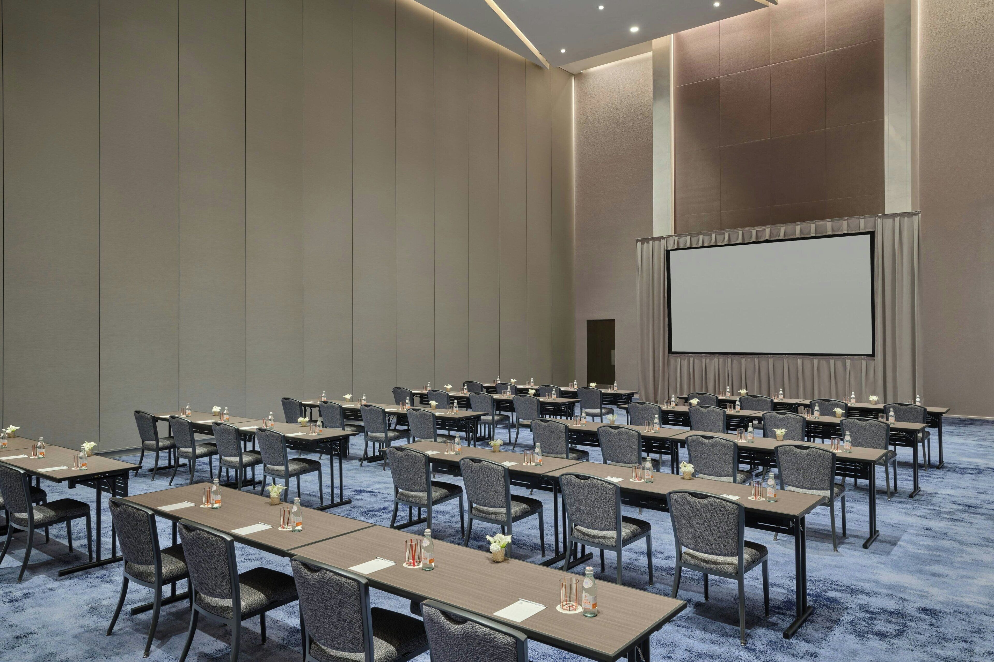 A conference room with rows of tables and chairs, a large screen, and a carpeted floor.