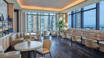 En modern hotellobby med högt i tak, stora fönster med utsikt över staden, bekväma sittgrupper och en väl upplyst atmosfär.