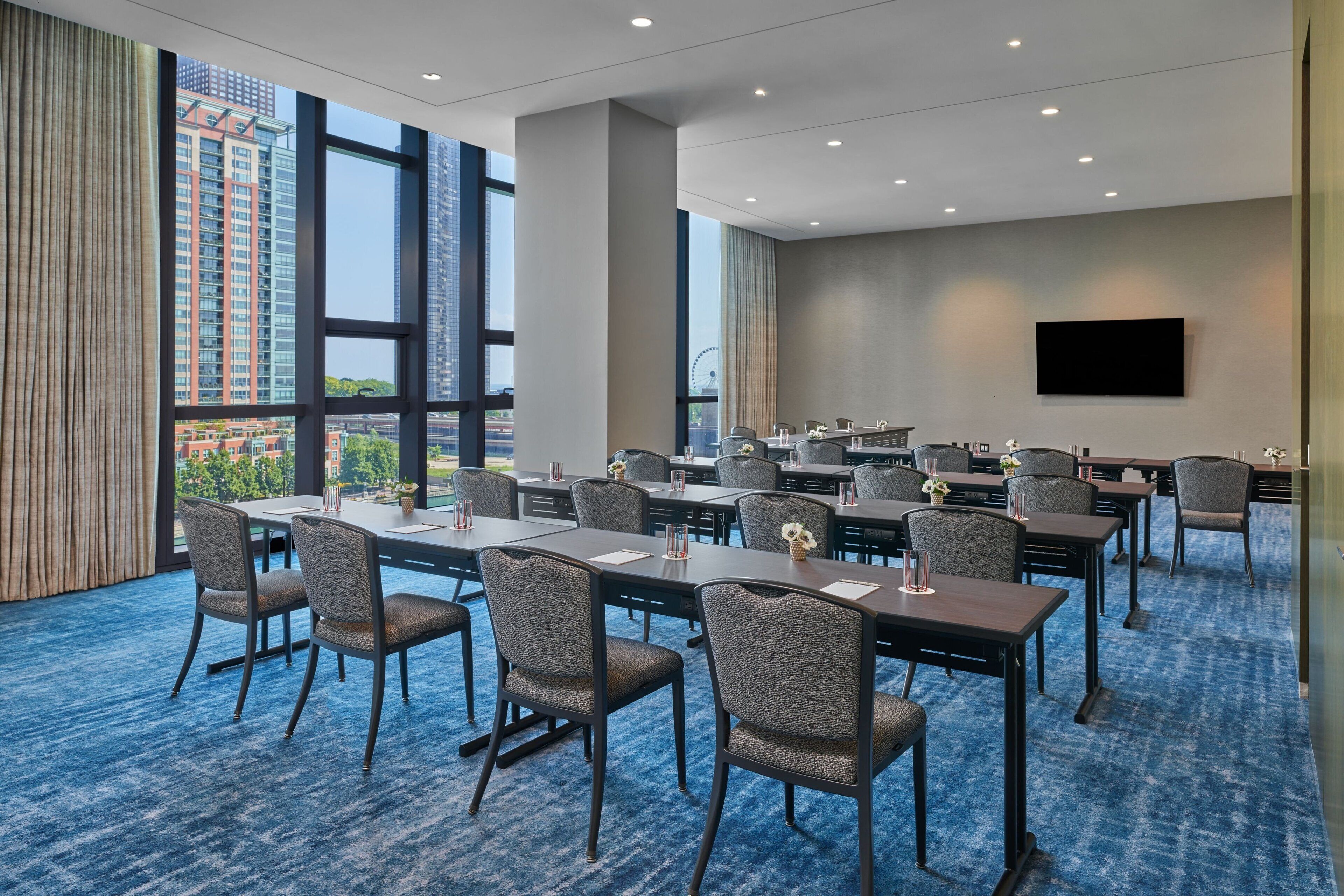 A conference room with tables and chairs, large windows, and a flat-screen TV on the wall.