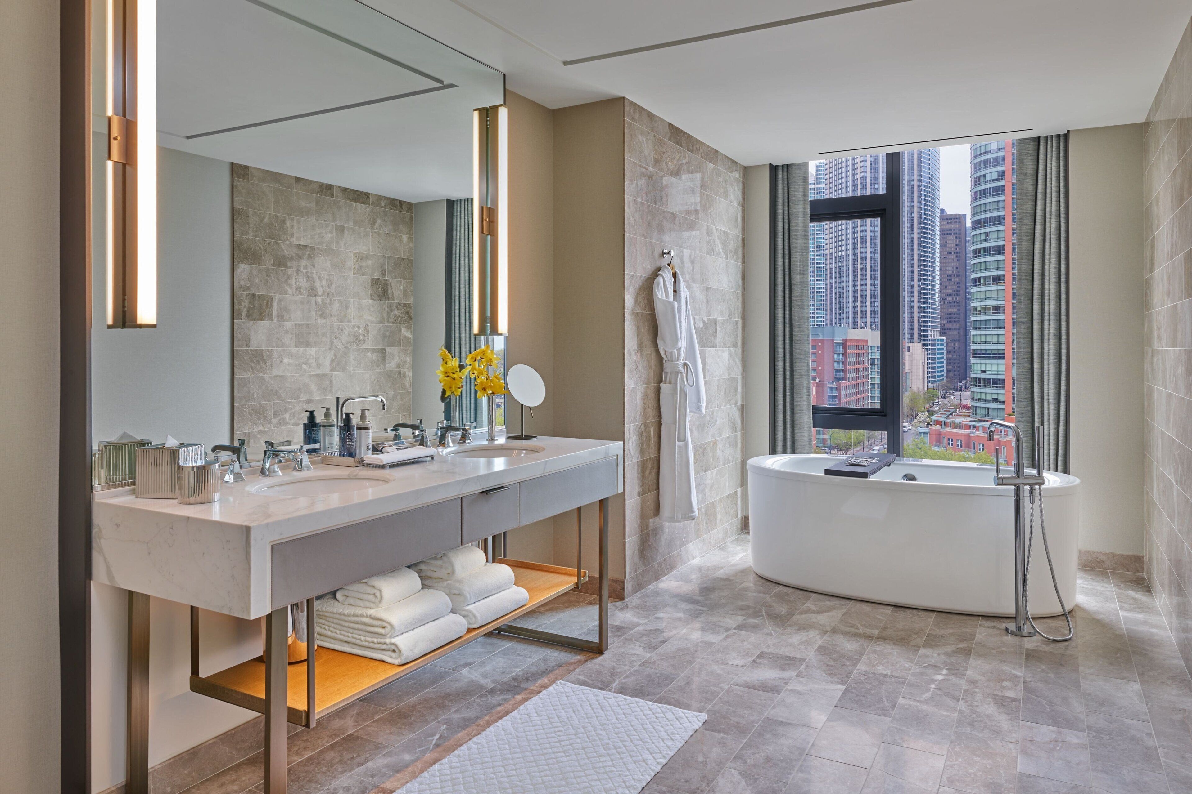 A modern bathroom with a large mirror, two sinks, a bathtub, and a view of the city.