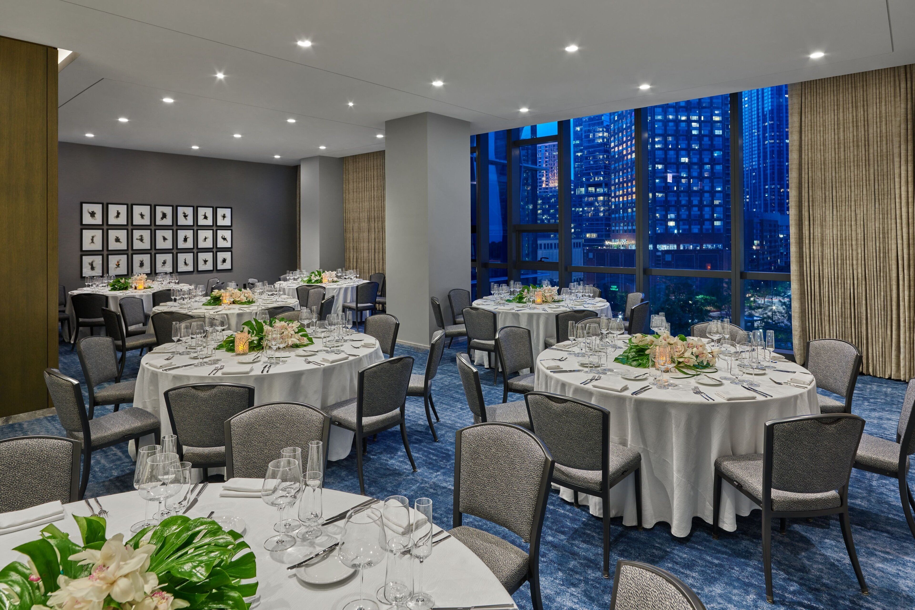 En hotellrestaurang med runda bord dukade för middag, stora fönster med utsikt över staden och inramade tavlor på väggen.
