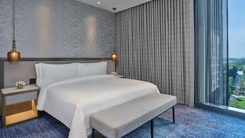 A modern hotel room with a large bed, a bench, a nightstand, and a view of the city through large windows.