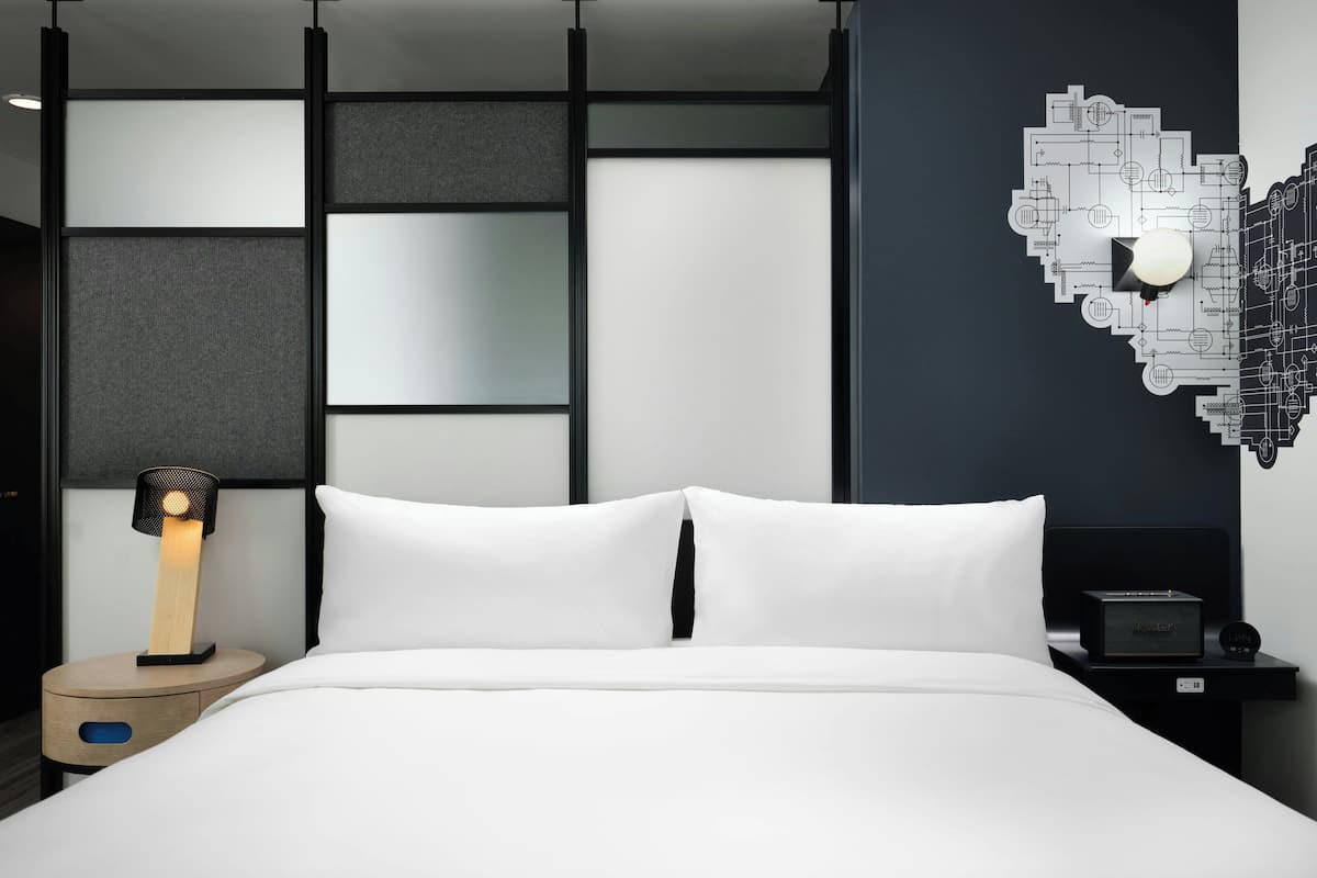 A modern hotel room with a bed, a nightstand with a lamp, a wall art piece, and a small table with a clock.