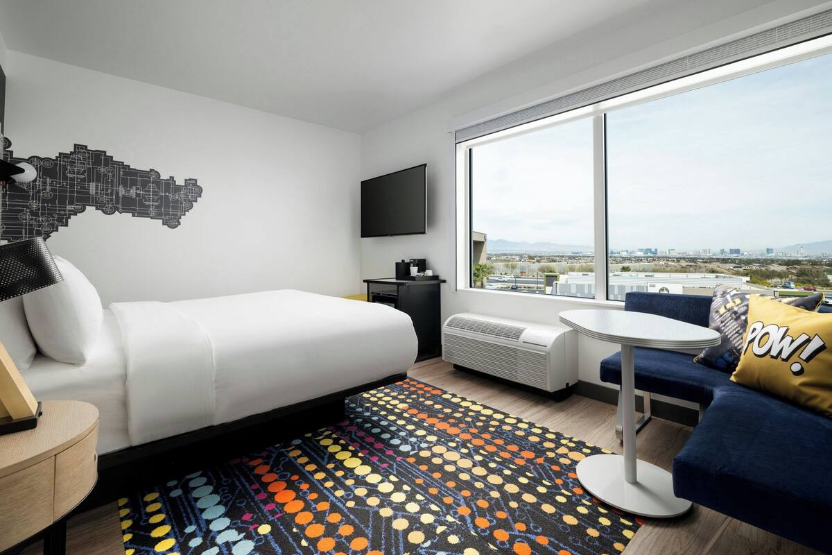 A modern hotel room with a bed, a sofa, a small table, a TV, and a large window offering a city view.