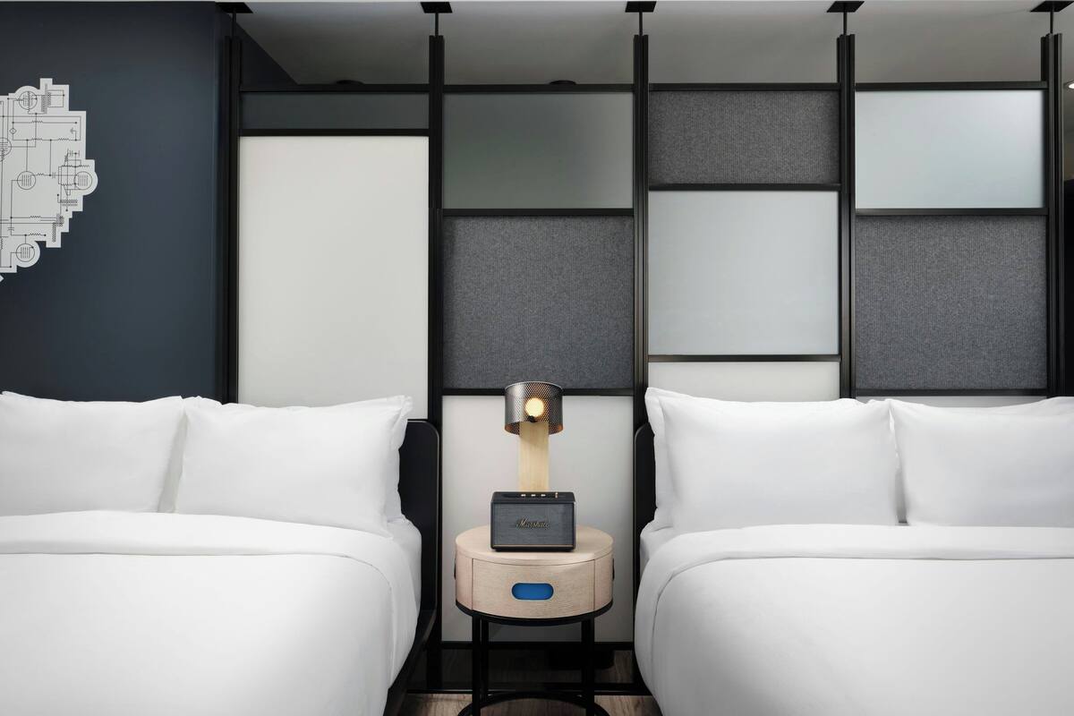 A modern hotel room with two beds, a nightstand with a lamp, and a wall-mounted control panel.