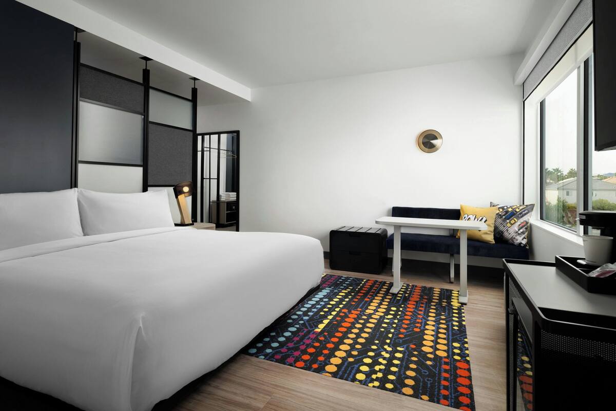 A modern hotel room with a large bed, a small desk, a sofa, and a view of the outdoors.
