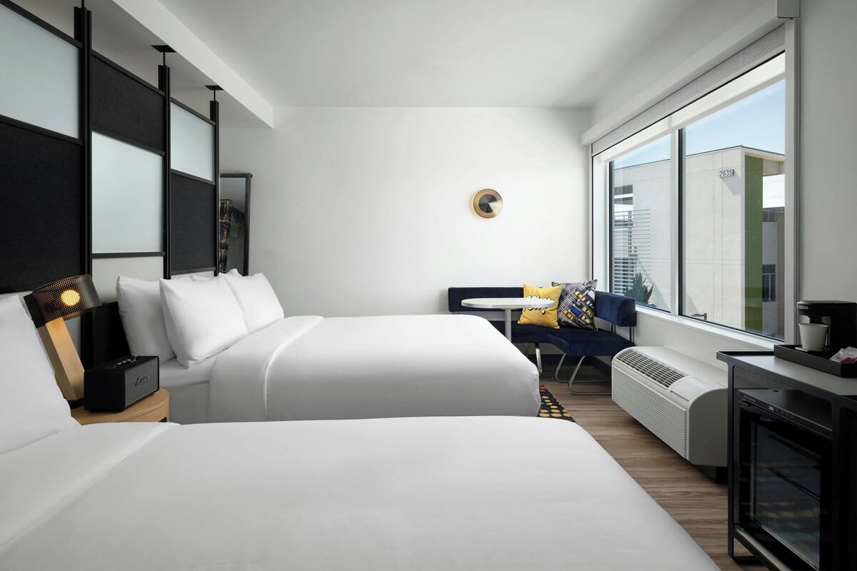 A modern hotel room with two beds, a desk, a chair, a heater, and a large window with a city view.