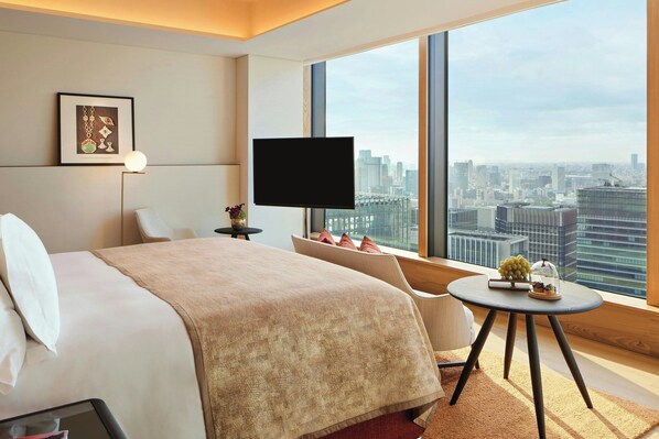 A hotel room with a large bed, a TV, a small table with a lamp, and a view of the city.