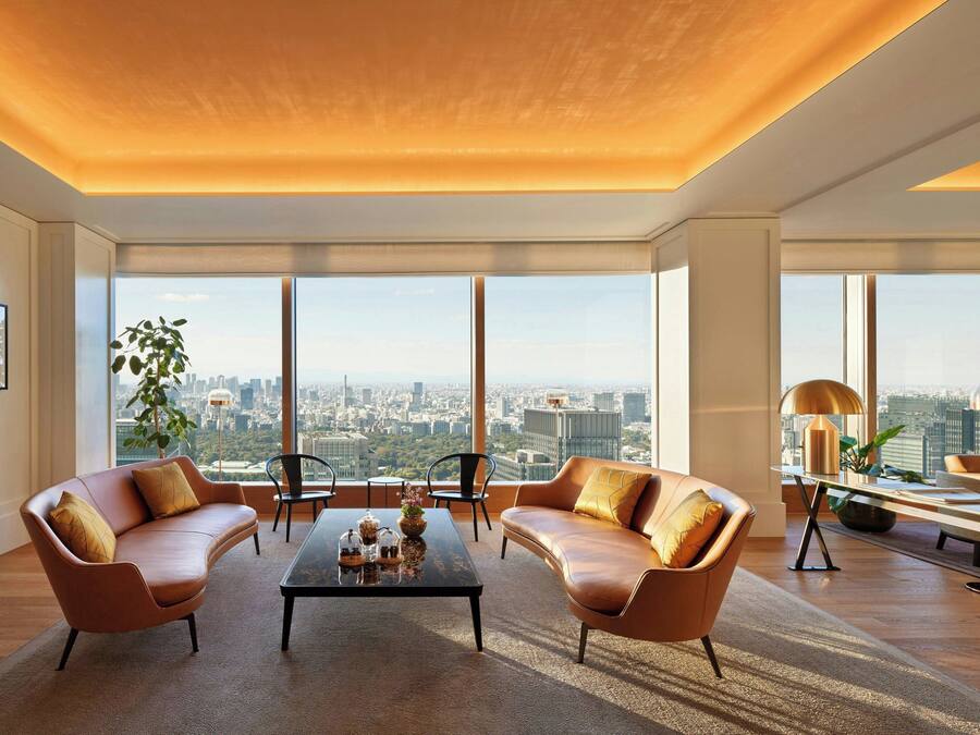 A modern living room with a city view, featuring two leather sofas, a coffee table, and a dining area with a lamp and plant.
