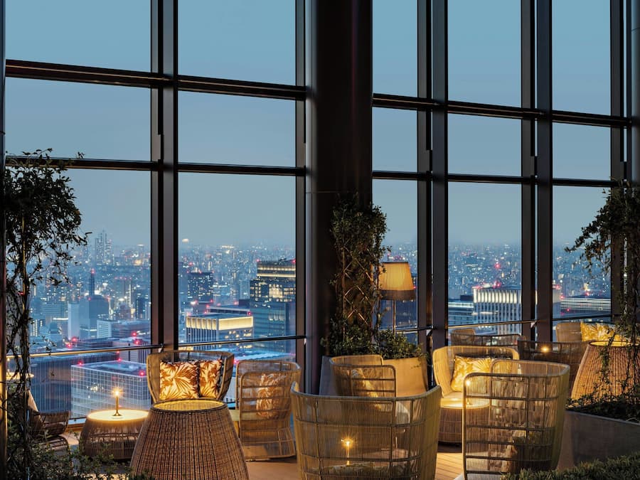 A high-rise hotel room with large windows offering a city view, wicker furniture, and a small table with a candle.