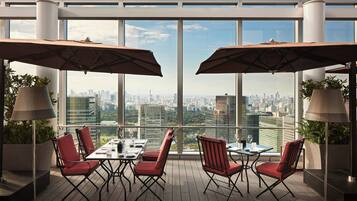 A rooftop terrace with a city view, furnished with red chairs and tables, and large umbrellas providing shade.