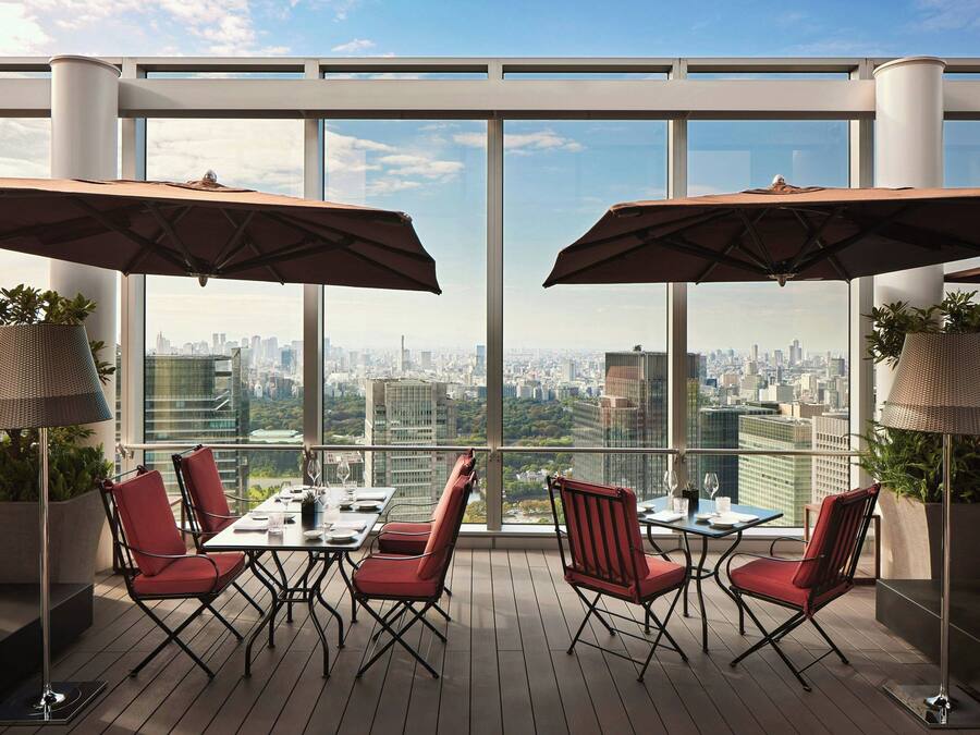 A rooftop terrace with a city view, furnished with red chairs and tables, and large umbrellas providing shade.