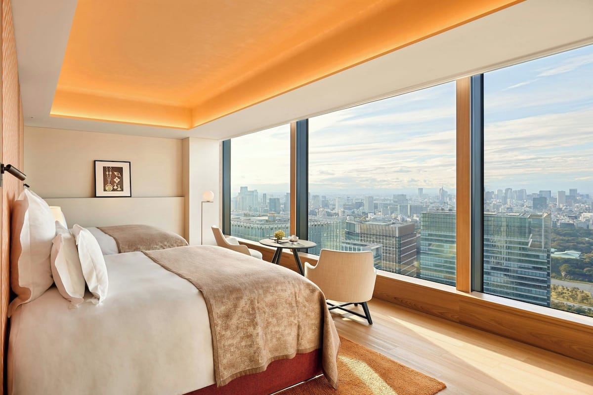 A modern hotel room with a large bed, a seating area, and a city view through large windows.