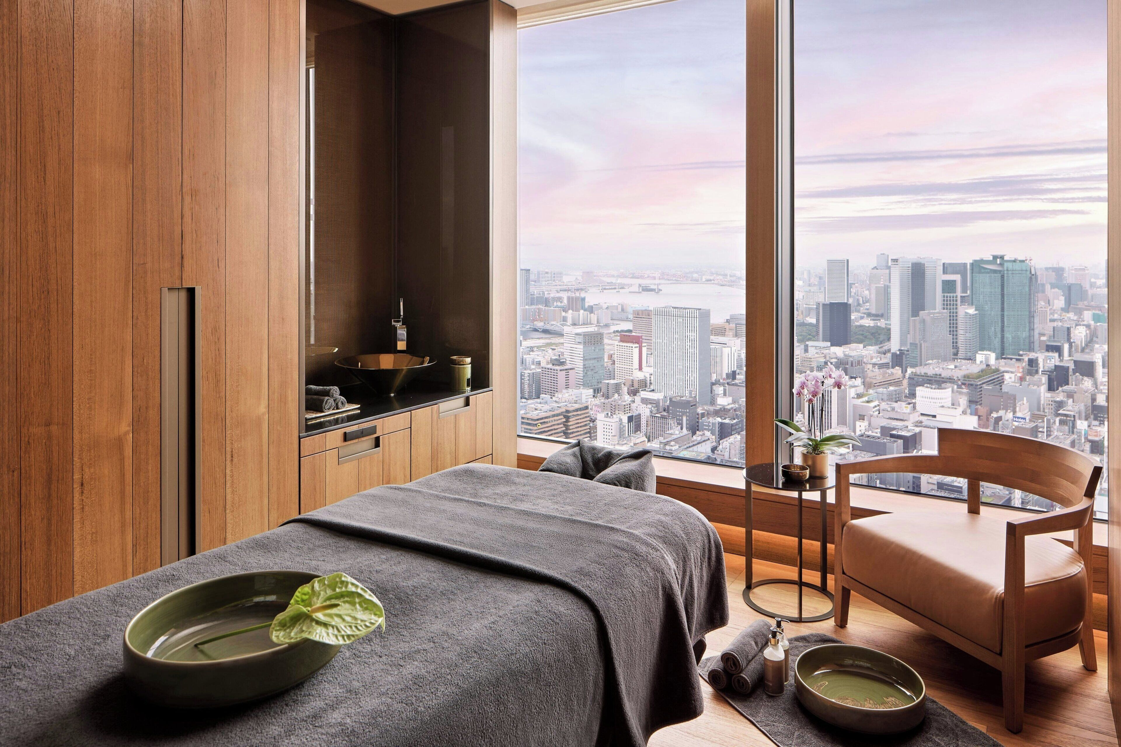 A modern room with a large window, wooden paneling, a massage table, a wooden chair, and a city view.