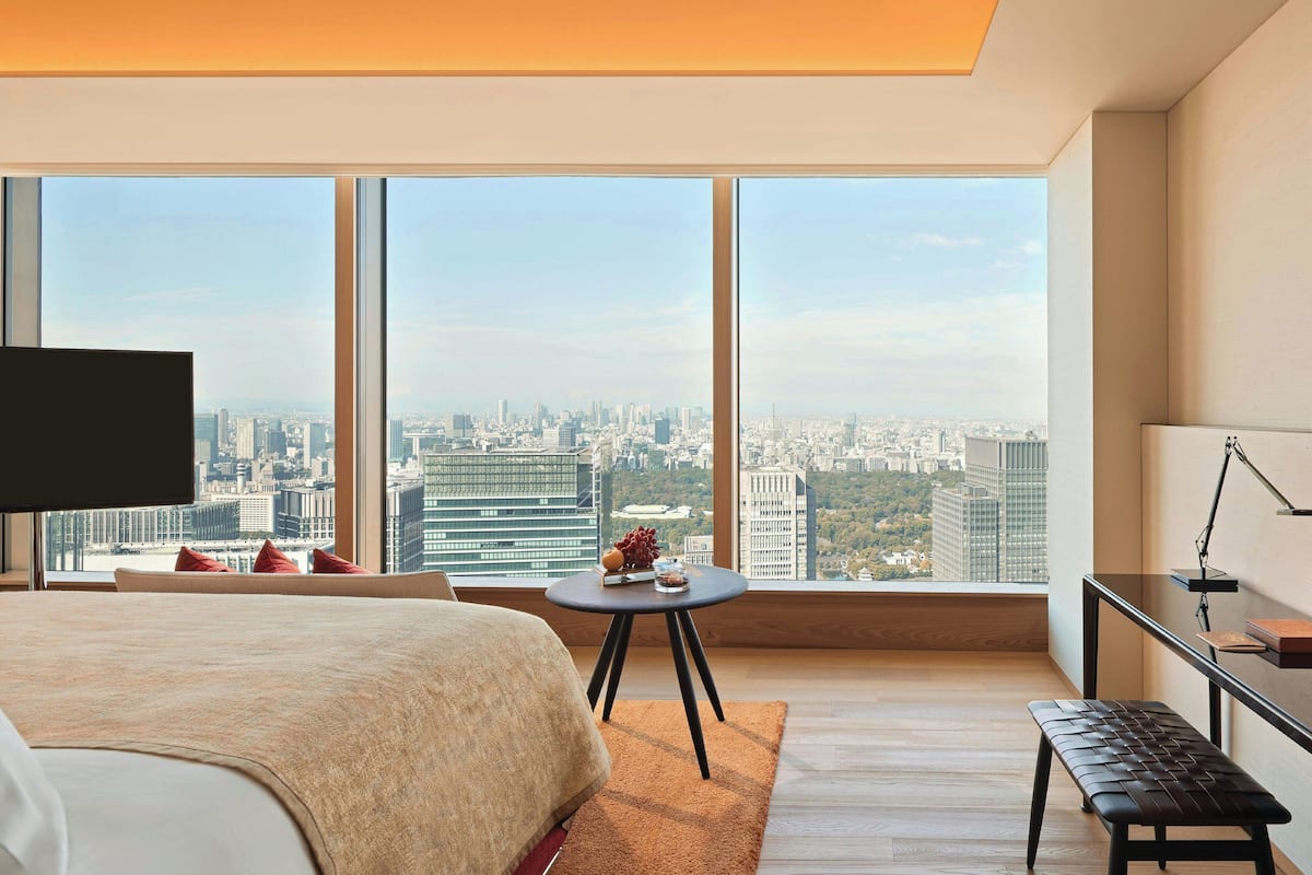 A modern hotel room with a large bed, a small round table, a bench, and a city view through large windows.