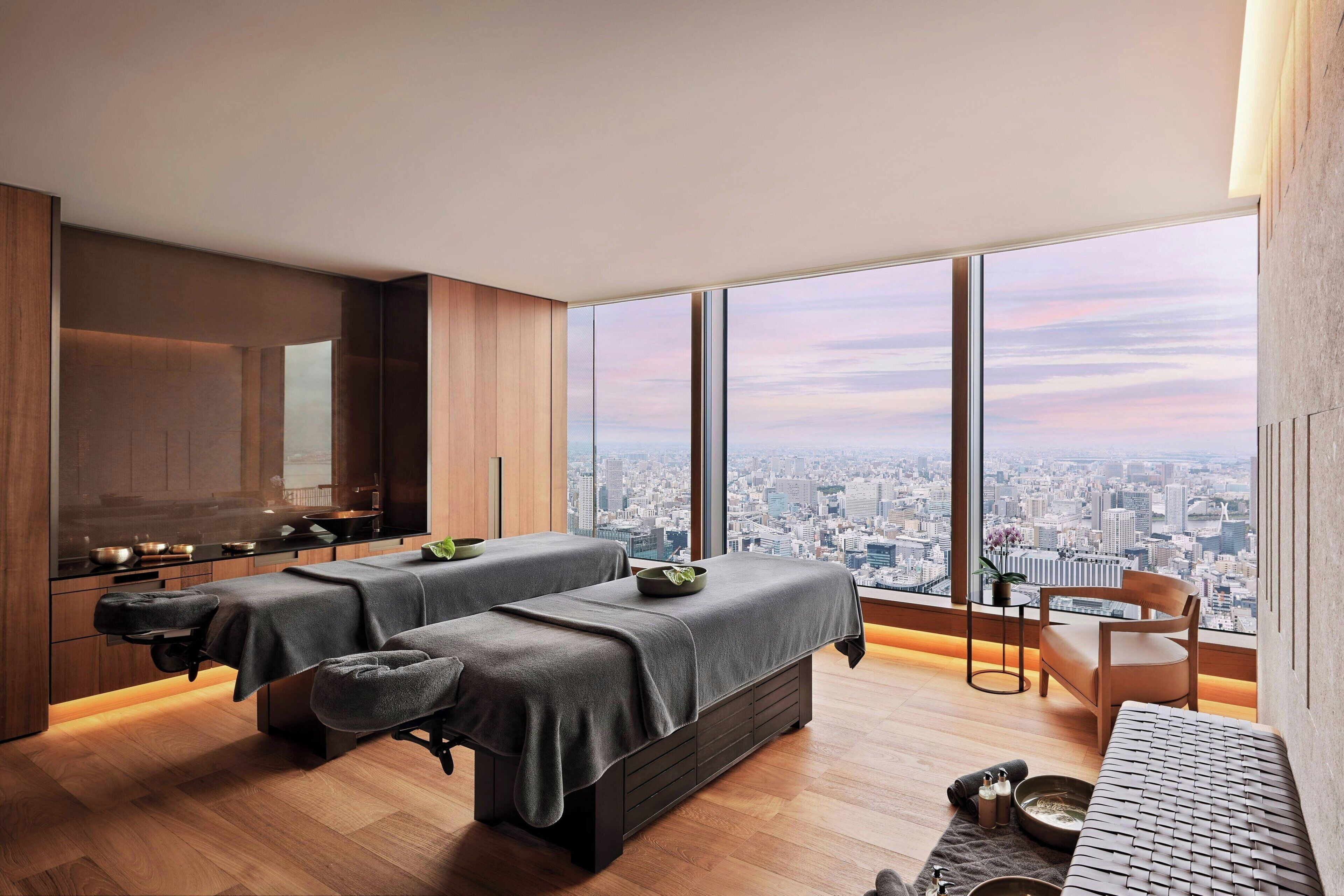 A modern spa room with two massage tables, a small seating area, and a city view through large windows.