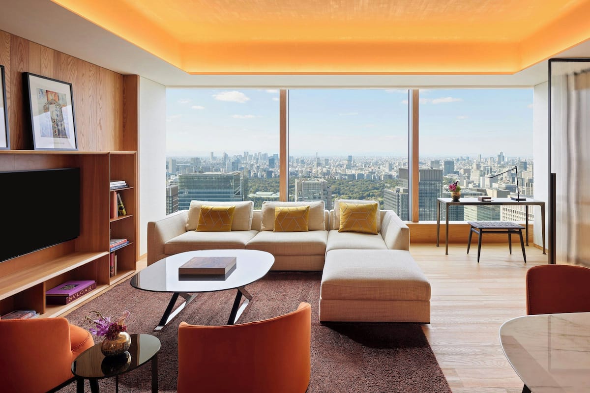 A modern living room with a large sofa, a coffee table, and a TV. There is a city view through large windows.