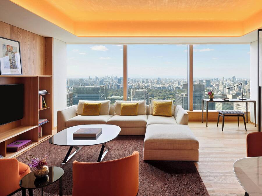 Una sala moderna con un sofá grande, una mesa de centro y un televisor. Se ve una vista urbana a través de amplias ventanas.