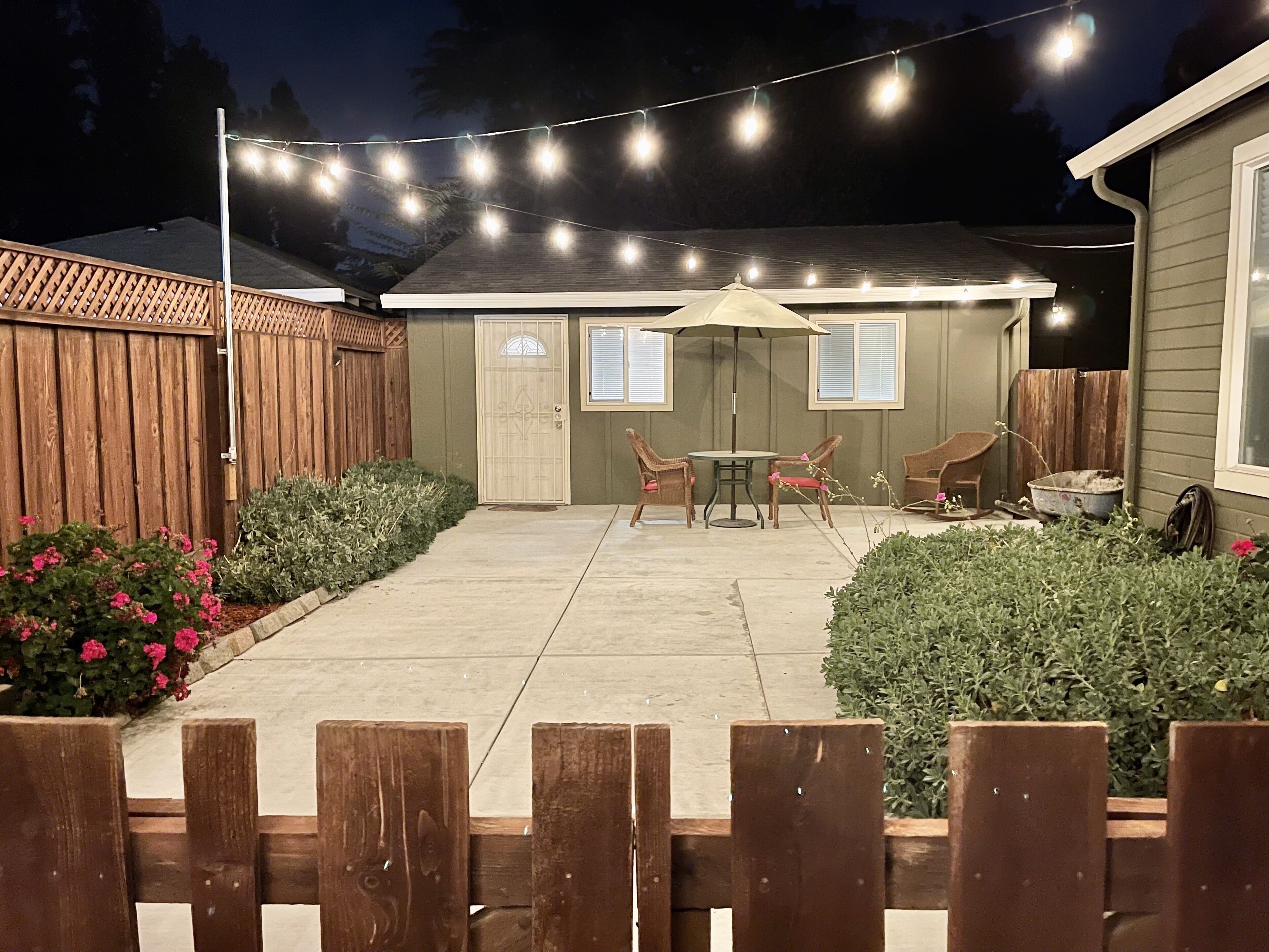 A backyard patio with a wooden fence, two chairs, a table, and string lights.