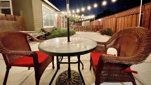 Un patio al aire libre con una mesa redonda y sillas de mimbre, un paraguas amarillo y luces colgantes.
