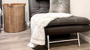 Una habitación de esquina con un sofá de cuero marrón, una manta blanca y un cojín gris. Hay una mesita de madera con una taza encima.