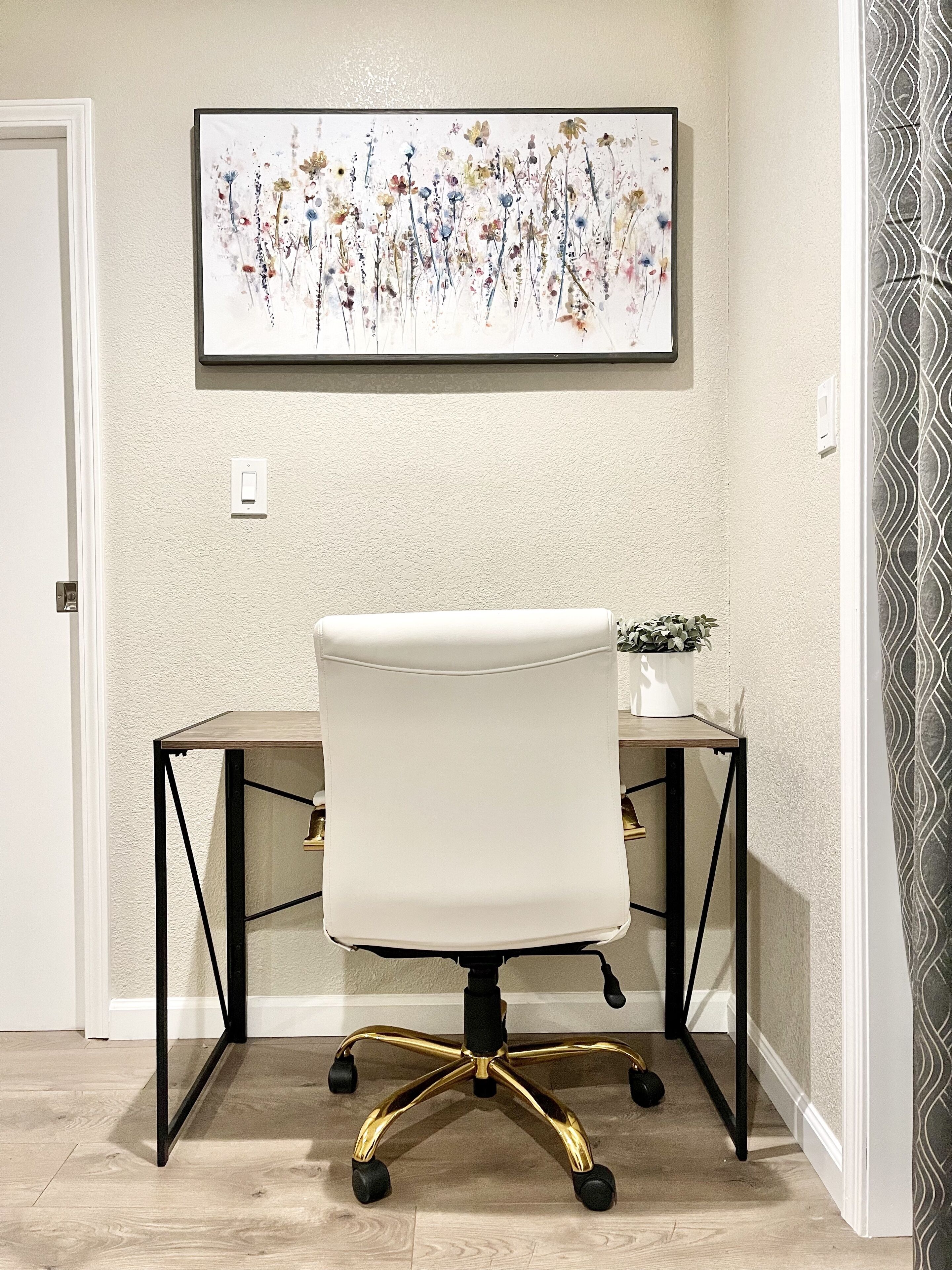 A white office chair with a black metal frame and gold accents on a desk, a framed floral artwork on the wall, and a patterned curtain.