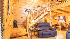 A wooden cabin interior with a staircase, a leather armchair, a dining table, and a chandelier. - Bearway to Heaven #2 (Pigeon Forge)