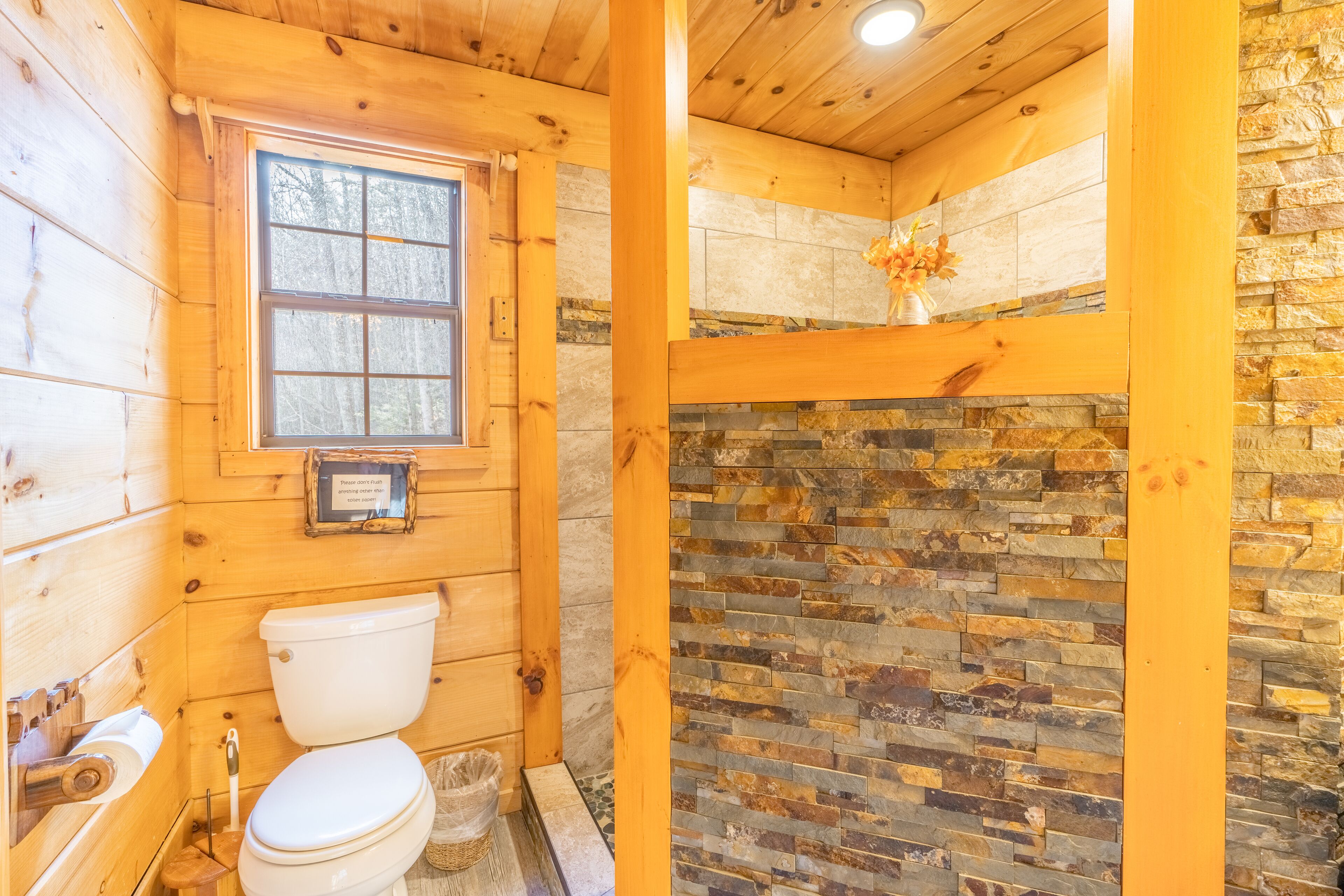 A wooden cabin bathroom with a stone wall, a window, and a toilet.