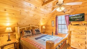 A cozy log cabin room with a bed, wooden headboard, a ceiling fan, a television, and a window with curtains. - Bearway to Heaven #2 (Pigeon Forge)