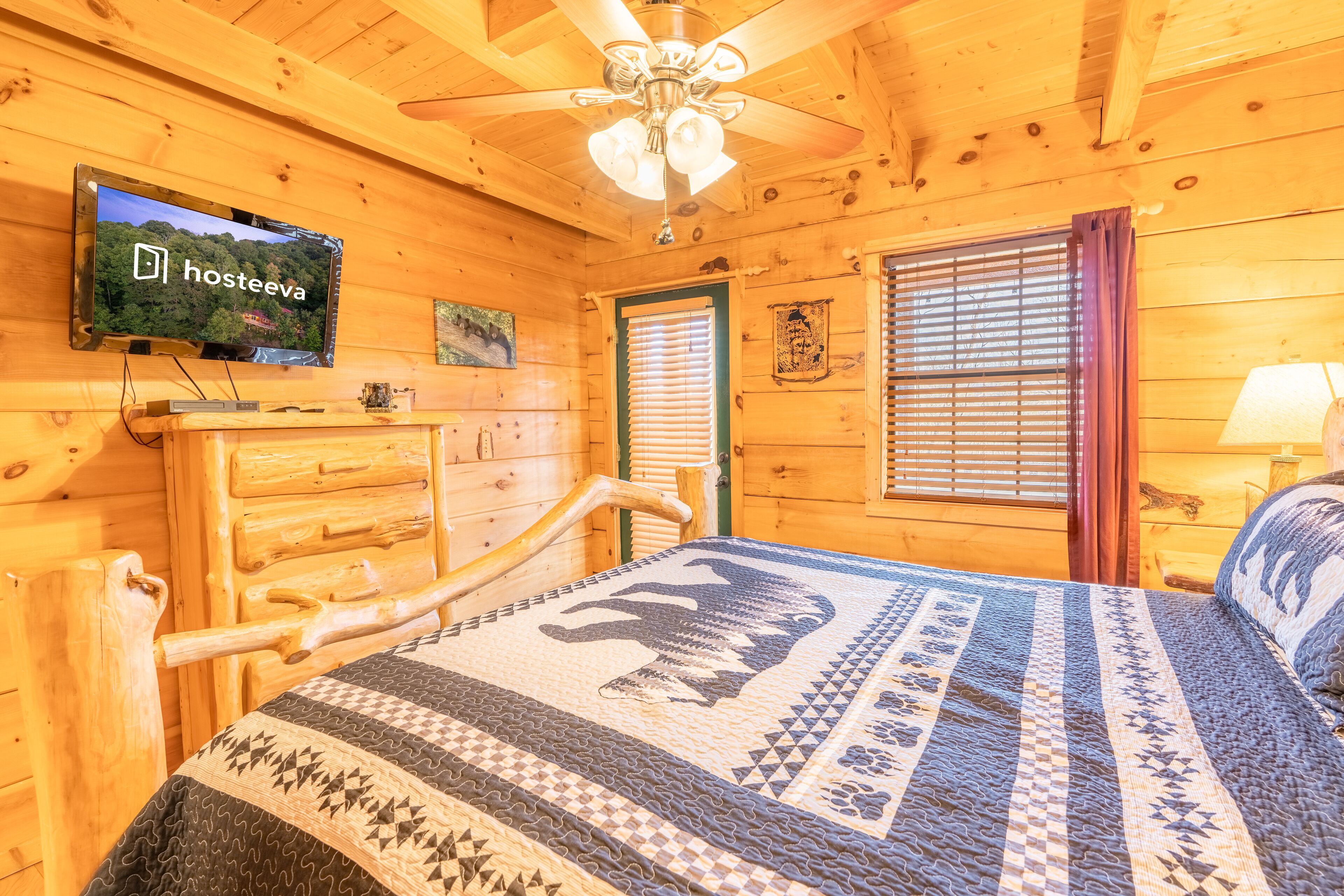 Ein Holzblockhaus-Zimmer mit einem Bett, einem an der Wand montierten Fernseher und einem Fenster mit Jalousien.