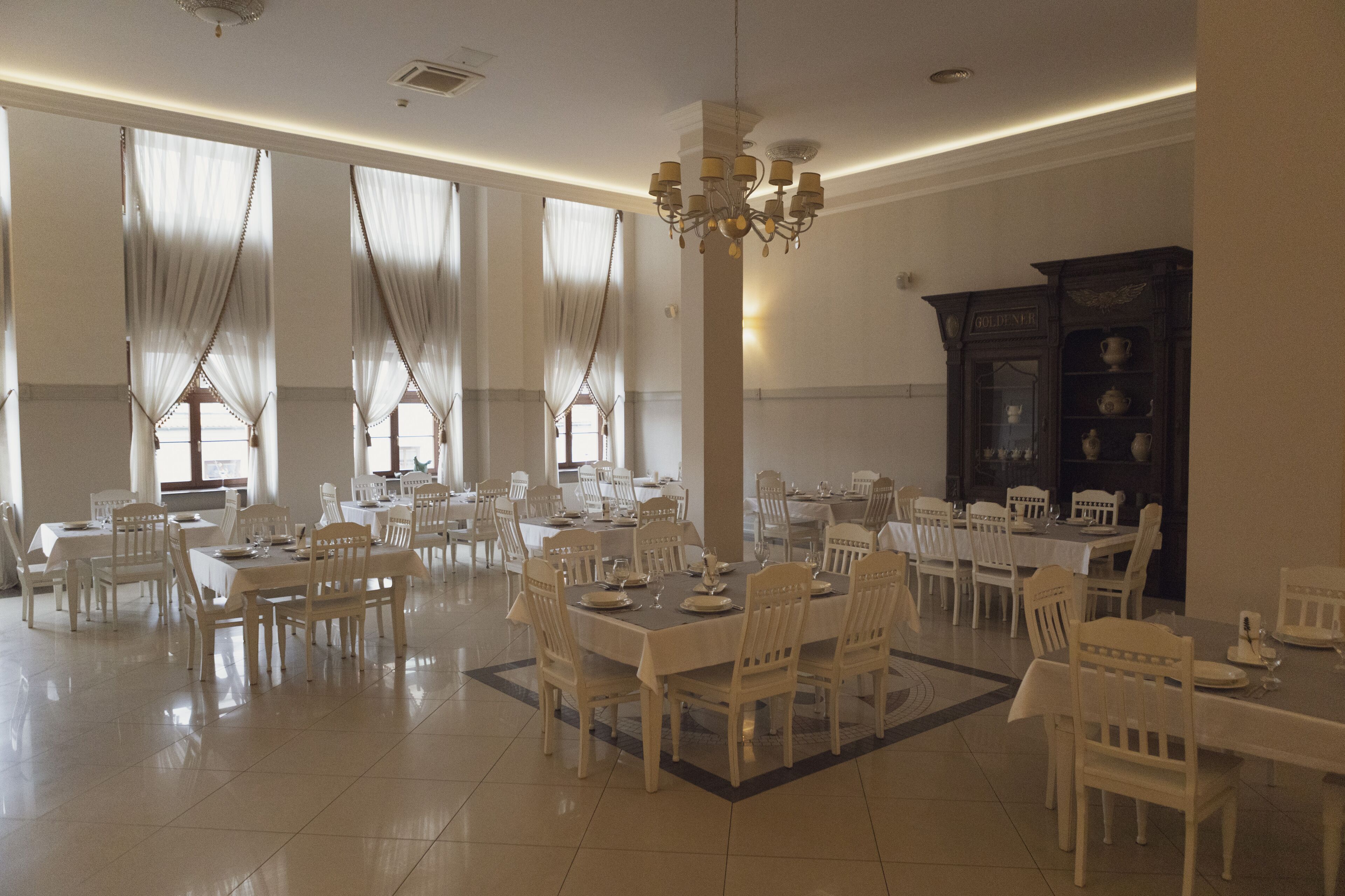 A spacious dining area with multiple tables and chairs, large windows with curtains, and a chandelier.
