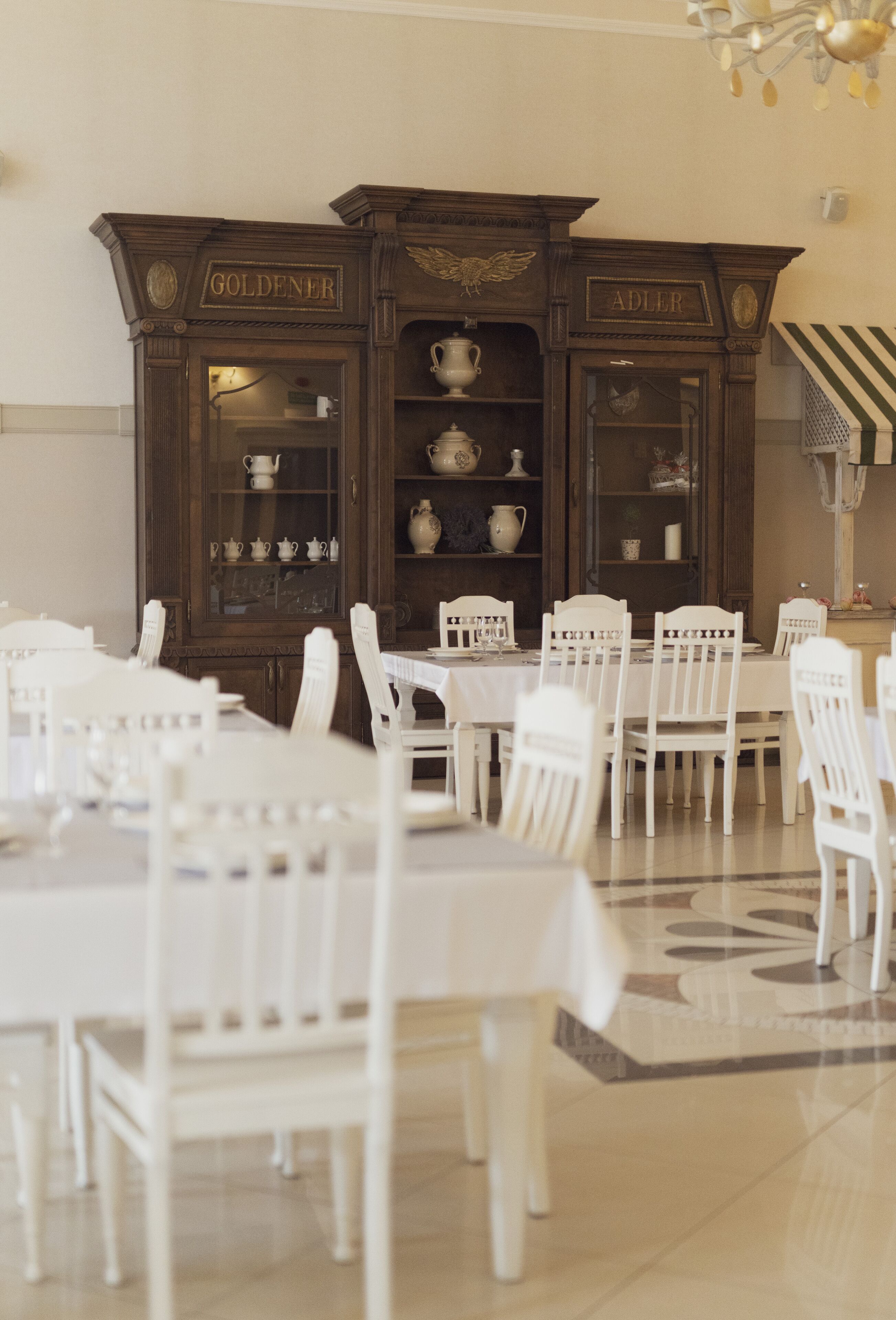 A dining area with white chairs and tables, a wooden cabinet with glass doors, and a chandelier.