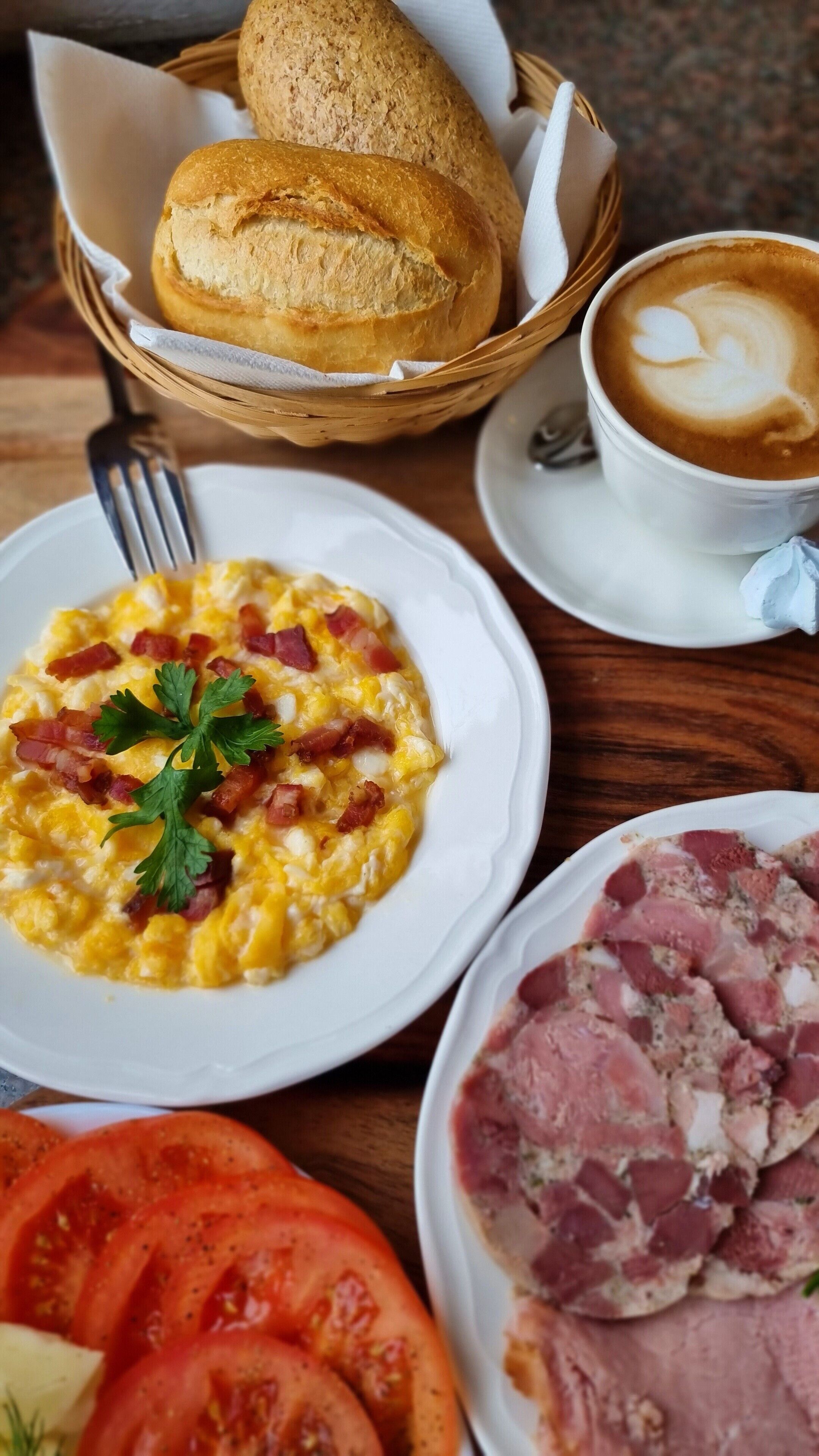 Mesa de café da manhã com ovos mexidos, bacon, presunto, tomates, pão e uma xícara de café.