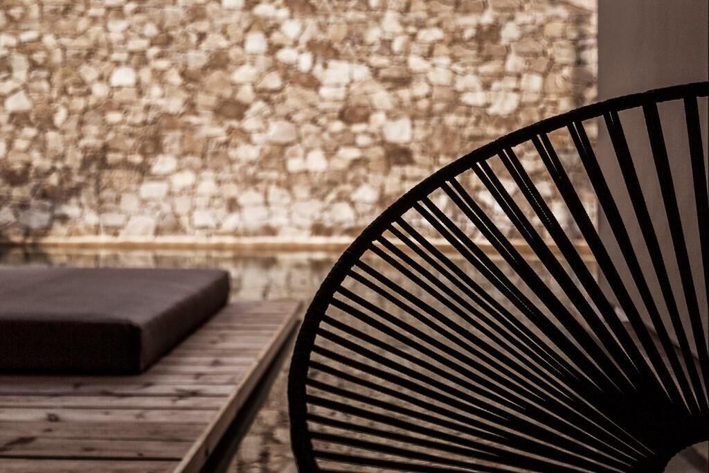 A wooden bench and a black chair with a radial design, positioned in front of a textured stone wall.