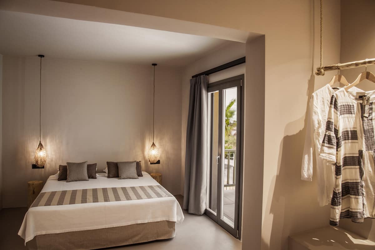 Une chambre moderne avec un grand lit, deux lampes suspendues, une armoire et une porte-fenêtre coulissante donnant sur l’extérieur.