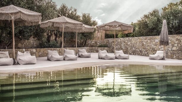 Zona de piscina con sillones de descanso y sombrillas, rodeada de muros de piedra y vegetación.