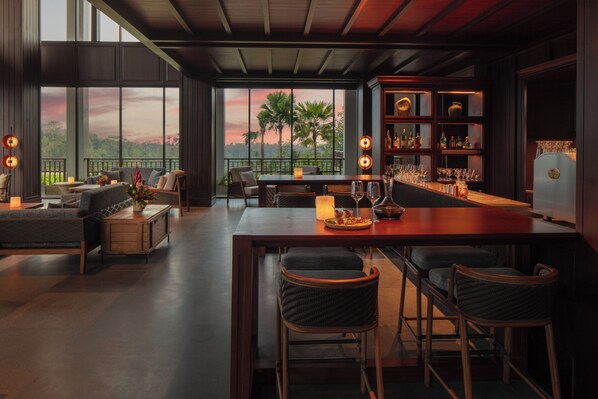 A spacious living area with a large window overlooking palm trees and a sunset, a bar with stools, and a bookshelf with various items. - Anantara Ubud Bali Resort (Payangan)