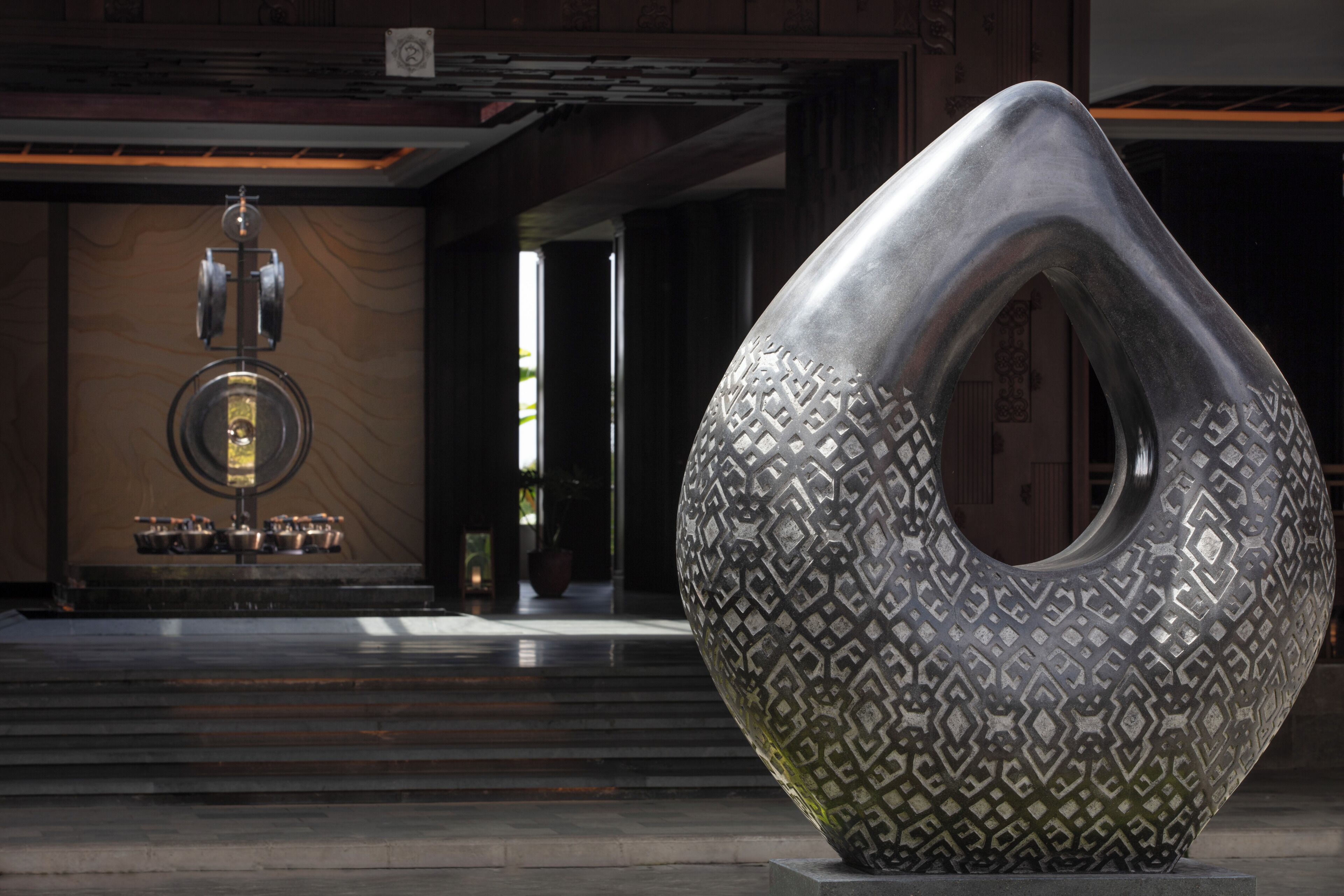 A modern sculpture in a lobby with a patterned design, a staircase, and a wall-mounted light fixture.
