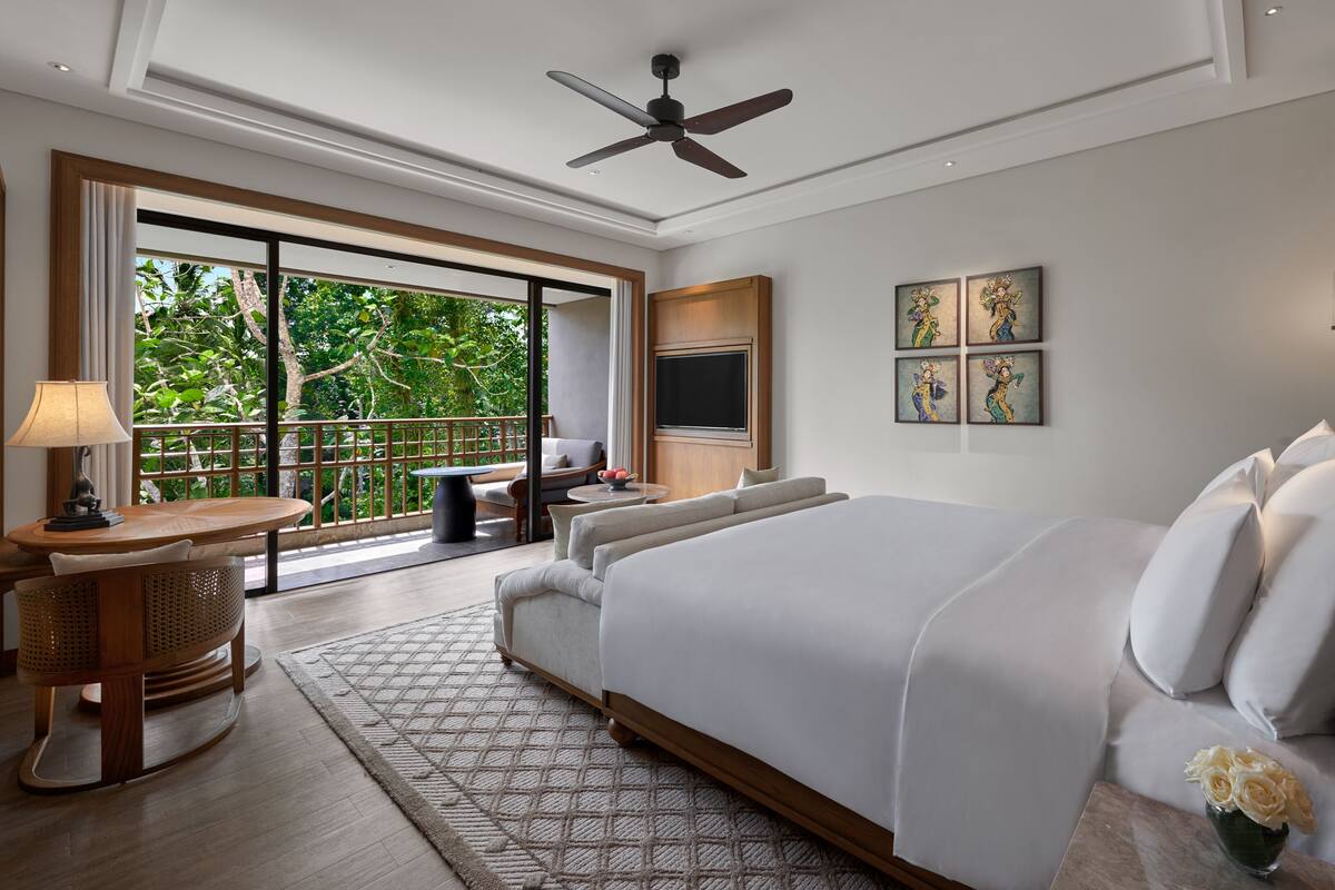 A spacious bedroom with a large bed, a balcony with a view of greenery, a ceiling fan, and artwork on the wall.
