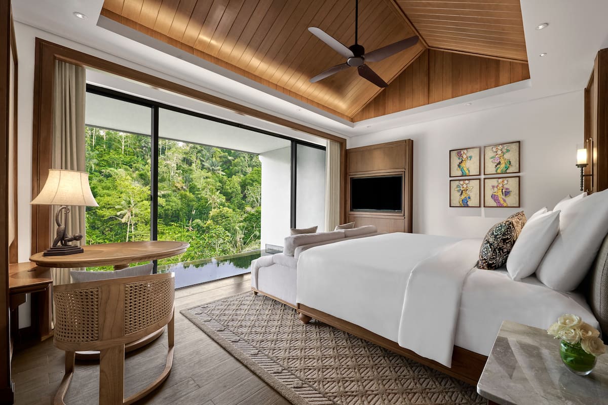 A spacious bedroom with a large bed, a ceiling fan, a TV, and a view of lush greenery.