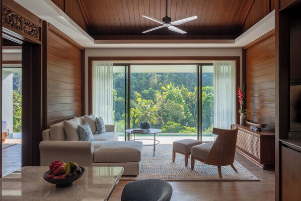 Una sala de estar moderna con un sofá, sillones, una mesa de centro y un televisor. La habitación tiene ventanas amplias con vistas a espacios verdes.