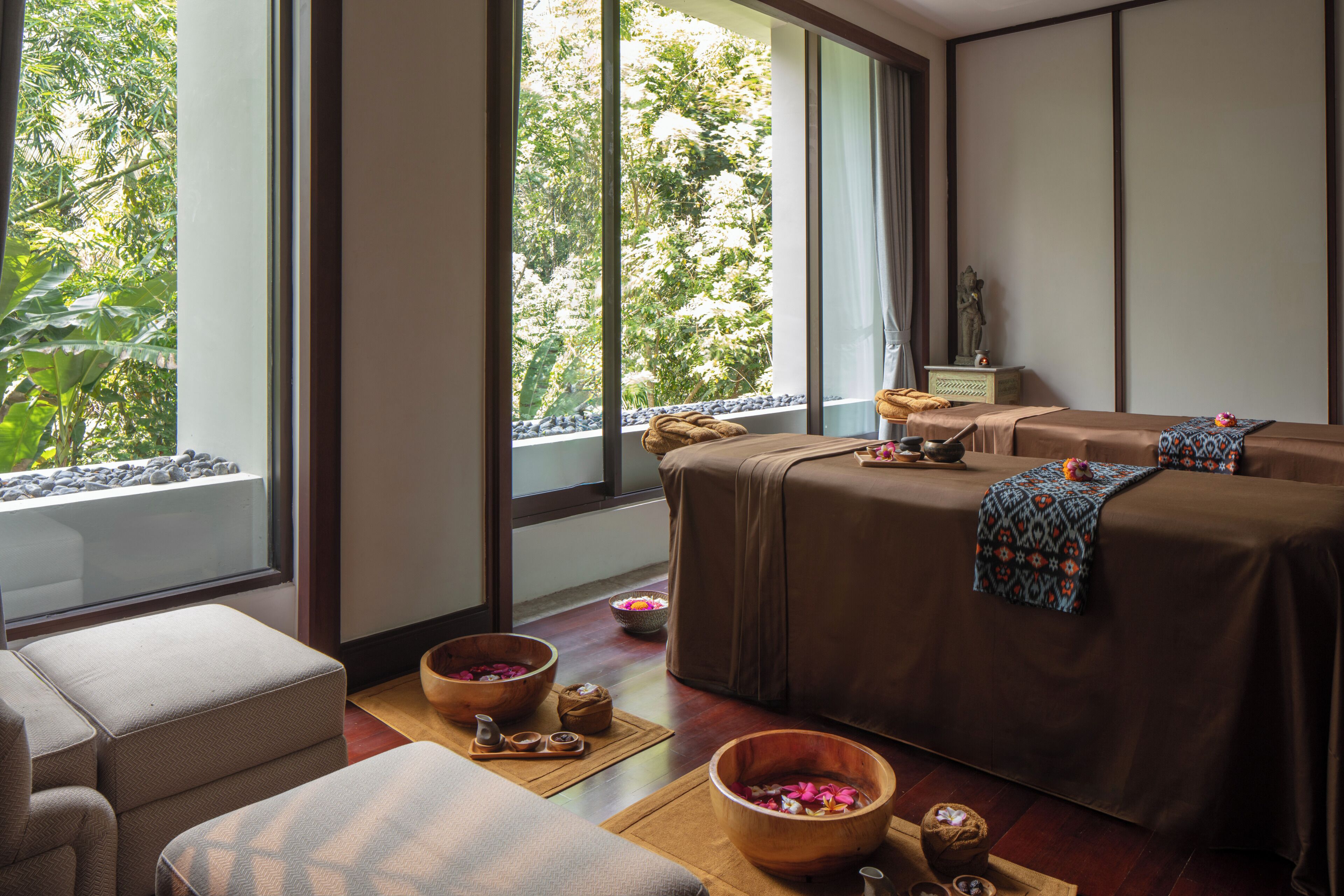 Un cuarto de spa con una camilla de masaje, un tazón de madera con flores y vista a árboles a través de amplios ventanales.
