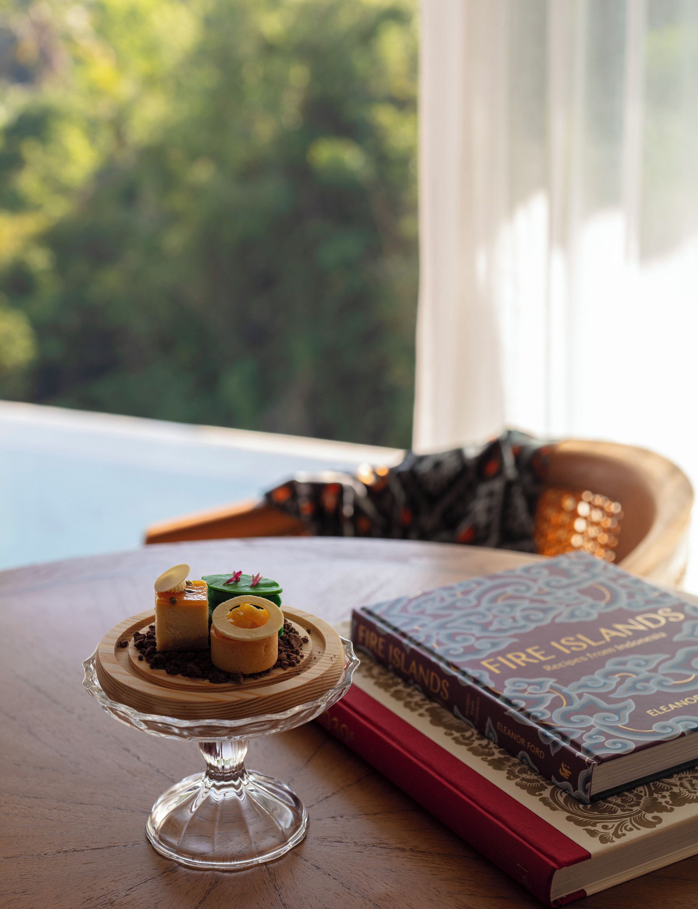 A dessert on a glass stand, a book titled "Fire Islands," and a view of greenery through a window.