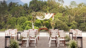 A wedding ceremony setup with a wooden altar, white chairs, and floral decorations on a wooden deck with a forest backdrop. - Anantara Ubud Bali Resort (Payangan)