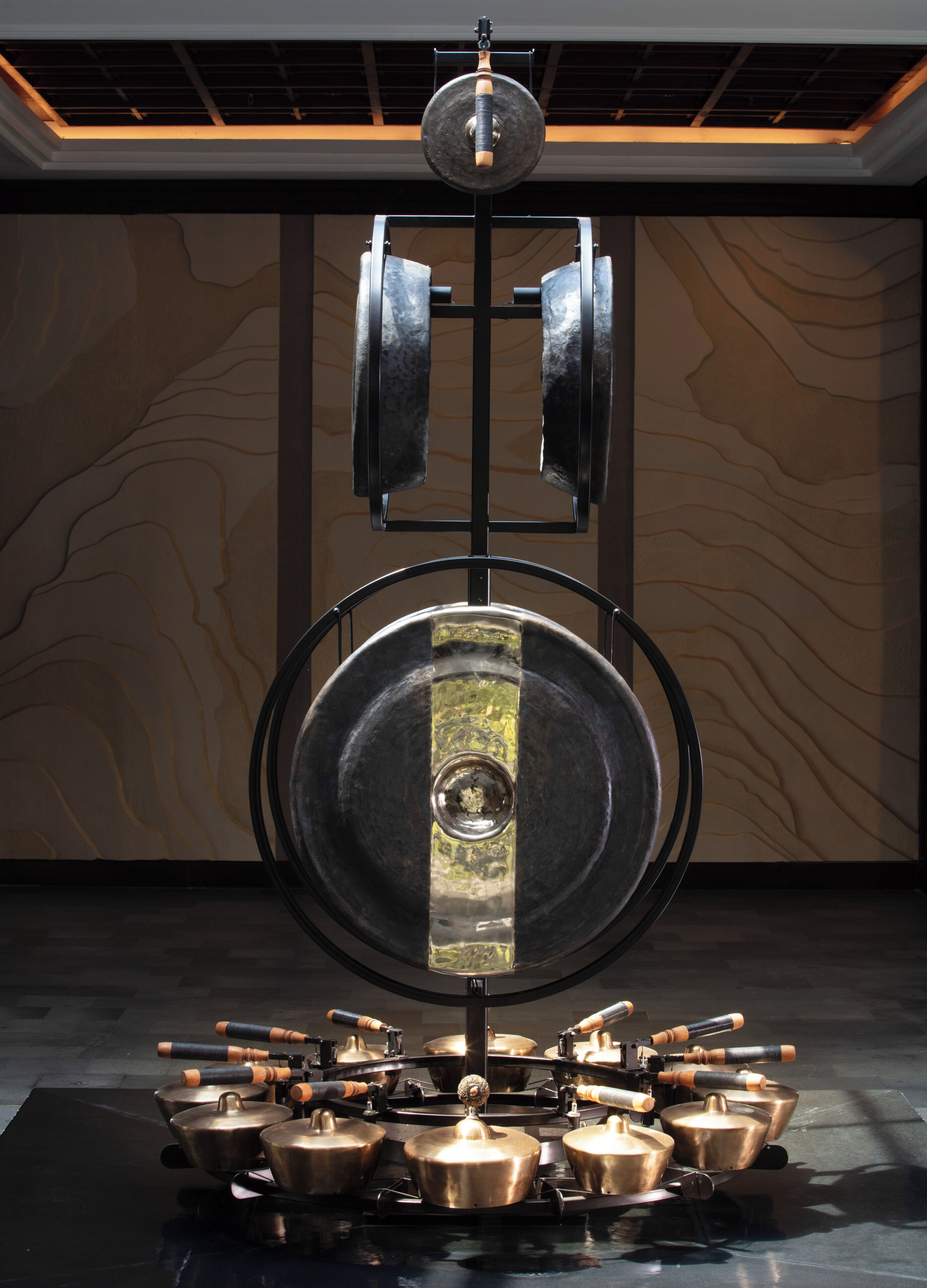 A large, ornate gong with a central bell, surrounded by a series of smaller bells and gong-like instruments on a reflective black surface.