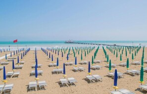 A beach with rows of sun loungers and umbrellas, a pier in the background, and a flag on the left. - Stylish Residenza TorreBianca 2 Bedroom Apartment sleeps 5 (Lignano Sabbiadoro)